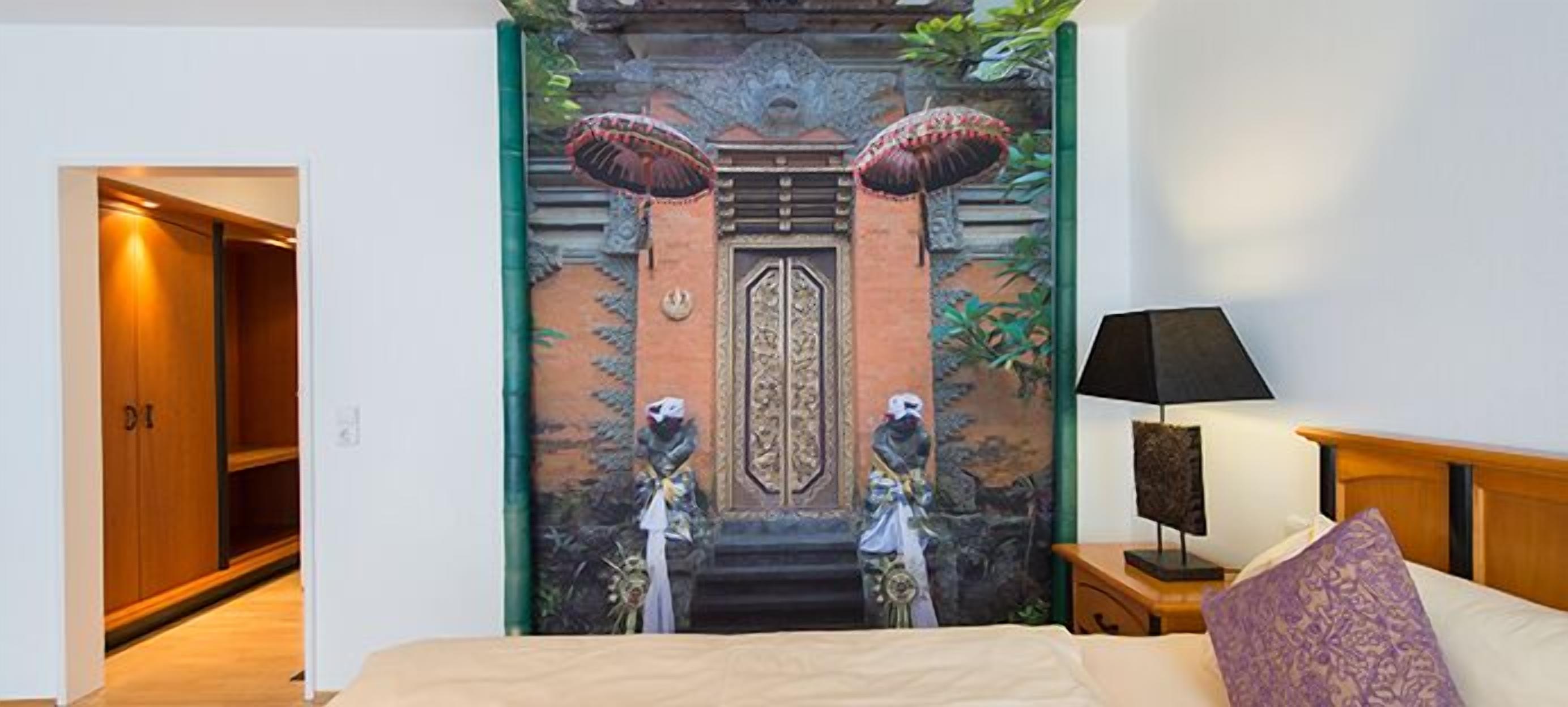 Ein Hotelzimmer mit Bett, Nachttisch, Lampe und einem Wandbild, das ein traditionelles Tor mit Personen zeigt.
