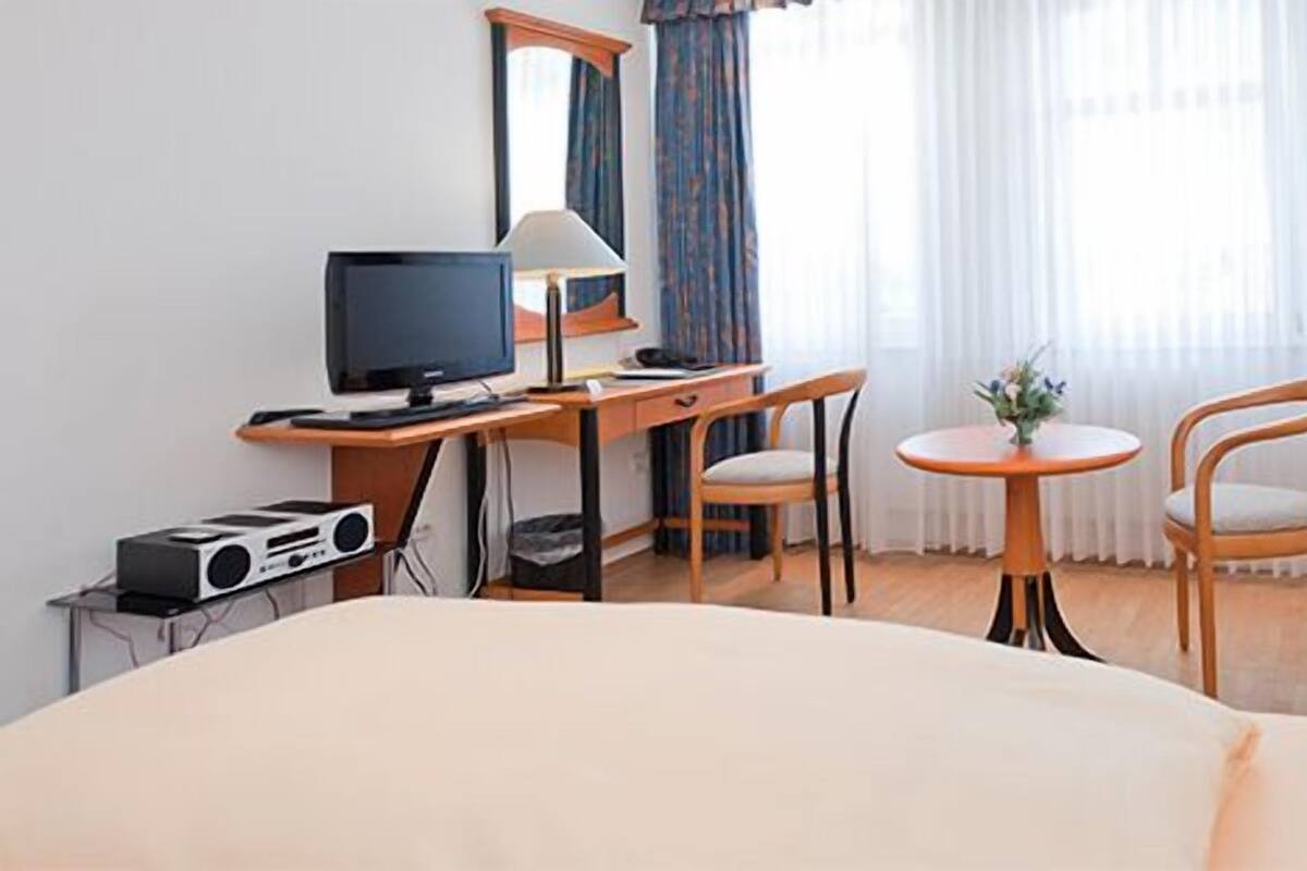 Ein Hotelzimmer mit Bett, Schreibtisch mit Computer, Stuhl, kleiner Tisch mit Lampe und Fernseher.