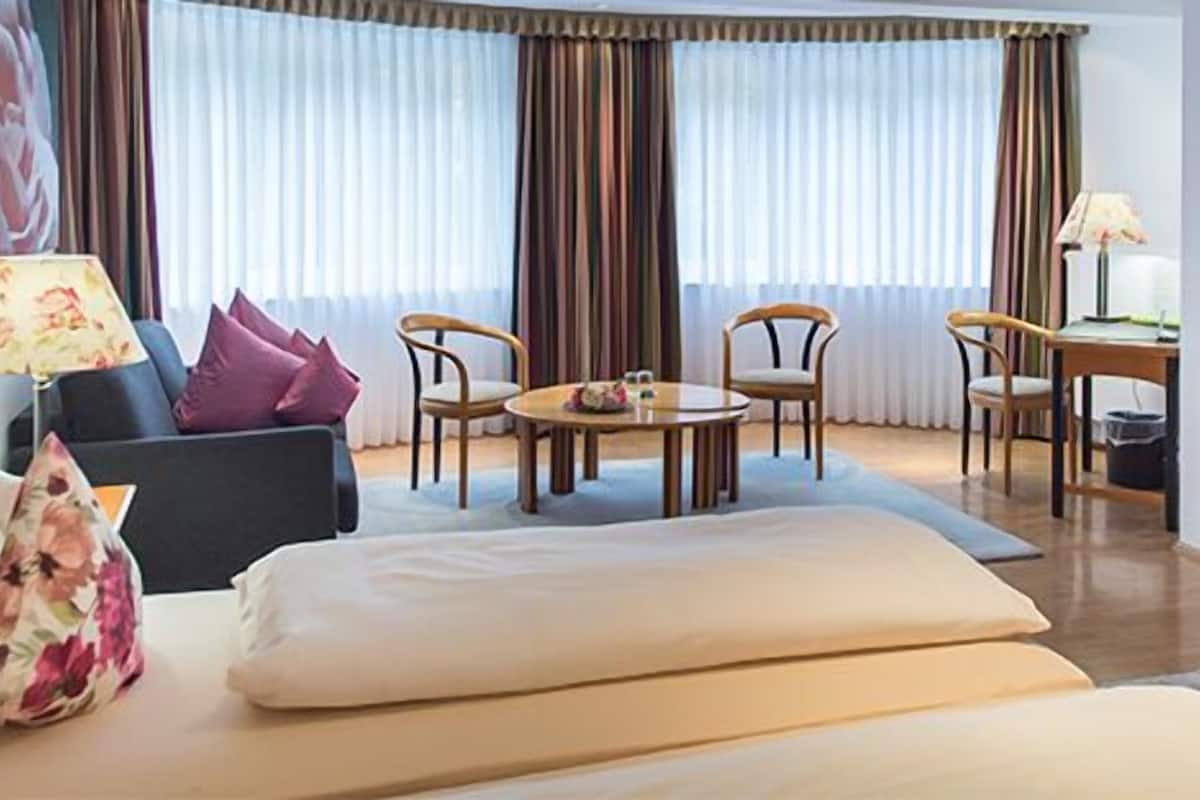 Ein Hotelzimmer mit einer Couch, zwei Sesseln, einem kleinen Tisch, einem Fernseher und einem großen Fenster mit Vorhängen.