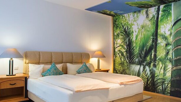 A double bed with two nightstands, each with a lamp, in a room with a tropical-themed mural on the wall. - Hotelpension Vitalis (Bad Hersfeld)