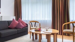 A modern living room with a grey sofa, two wooden chairs, a round wooden table, and a large window with curtains. - Hotelpension Vitalis (Bad Hersfeld)