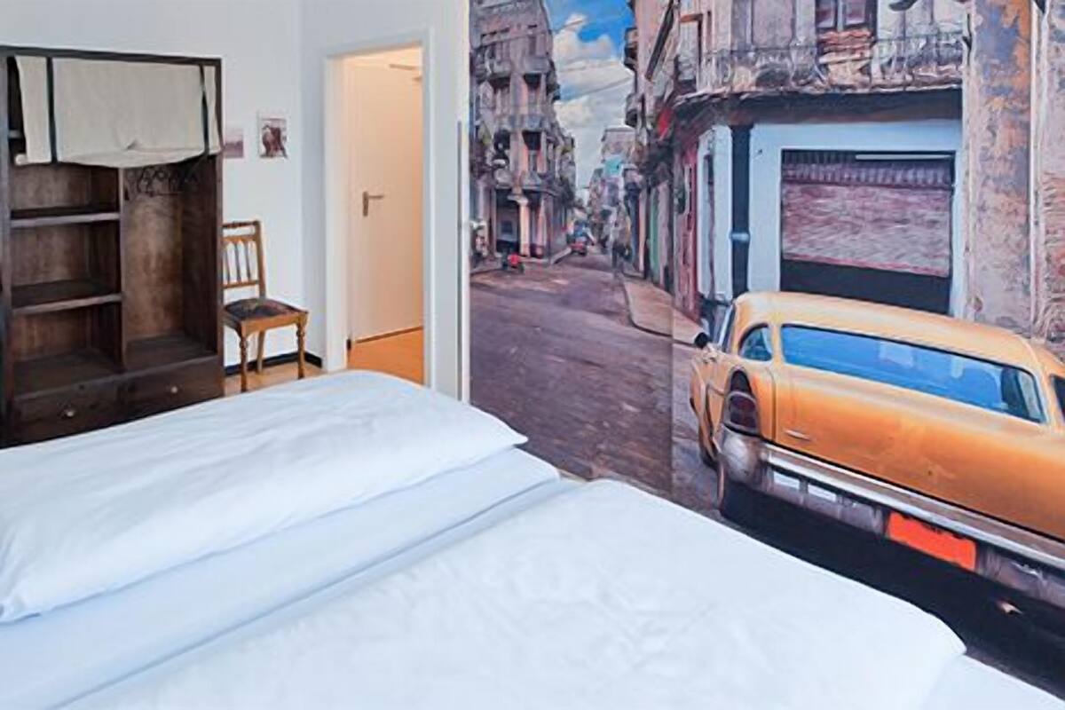Ein Hotelzimmer mit Bett, Schreibtisch, Stuhl, Kleiderschrank und einem Wandgemälde einer Straßenszene.