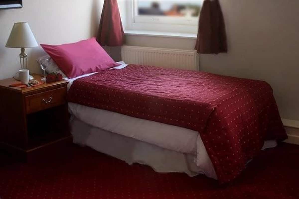 A neatly made bed with a red bedspread, a bedside table with a lamp, and a window with curtains.