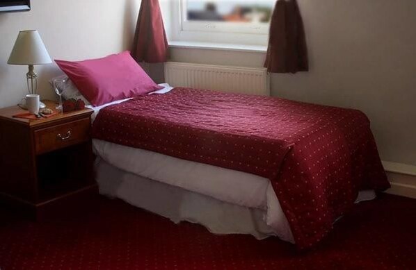 A neatly made bed with a red bedspread, a bedside table with a lamp, and a window with curtains. - The Griffin Hotel (Attleborough)