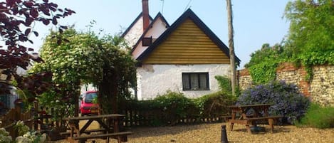 Una casa rústica con techo a dos aguas, una chimenea y una ventana. Se observan mesas de picnic y un coche rojo estacionado al fondo.