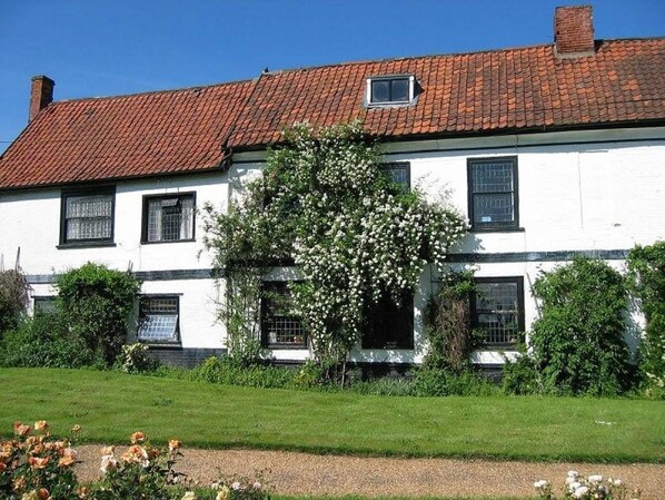 Un bâtiment blanc à deux étages, surmonté d’un toit recouvert de tuiles rouges, et entouré d’un jardin bien entretenu où poussent des rosiers et des arbustes.