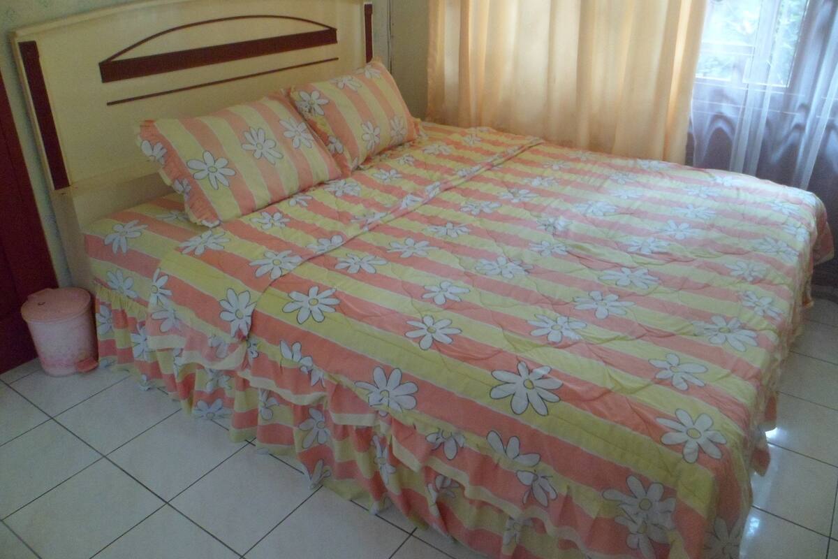 A bed with a floral-patterned bedspread, a wooden headboard, and a pink trash bin beside it.