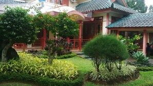 A courtyard with a well-maintained garden, a small tree, and a building with a tiled roof and red accents. - Kota Bunga BB (Cipanas)