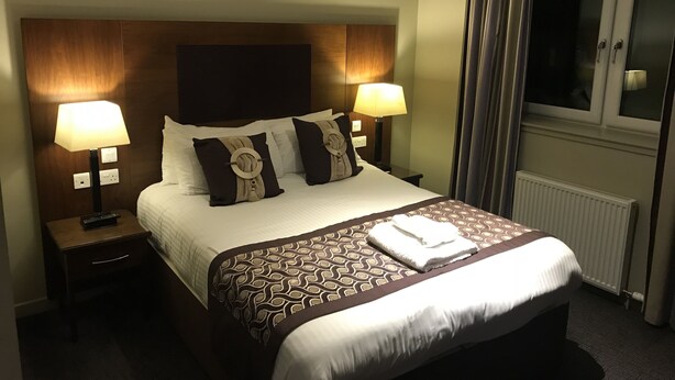 A hotel room with a large bed, two bedside lamps, a window with curtains, and a radiator. - The Buchan Hotel (Ellon)