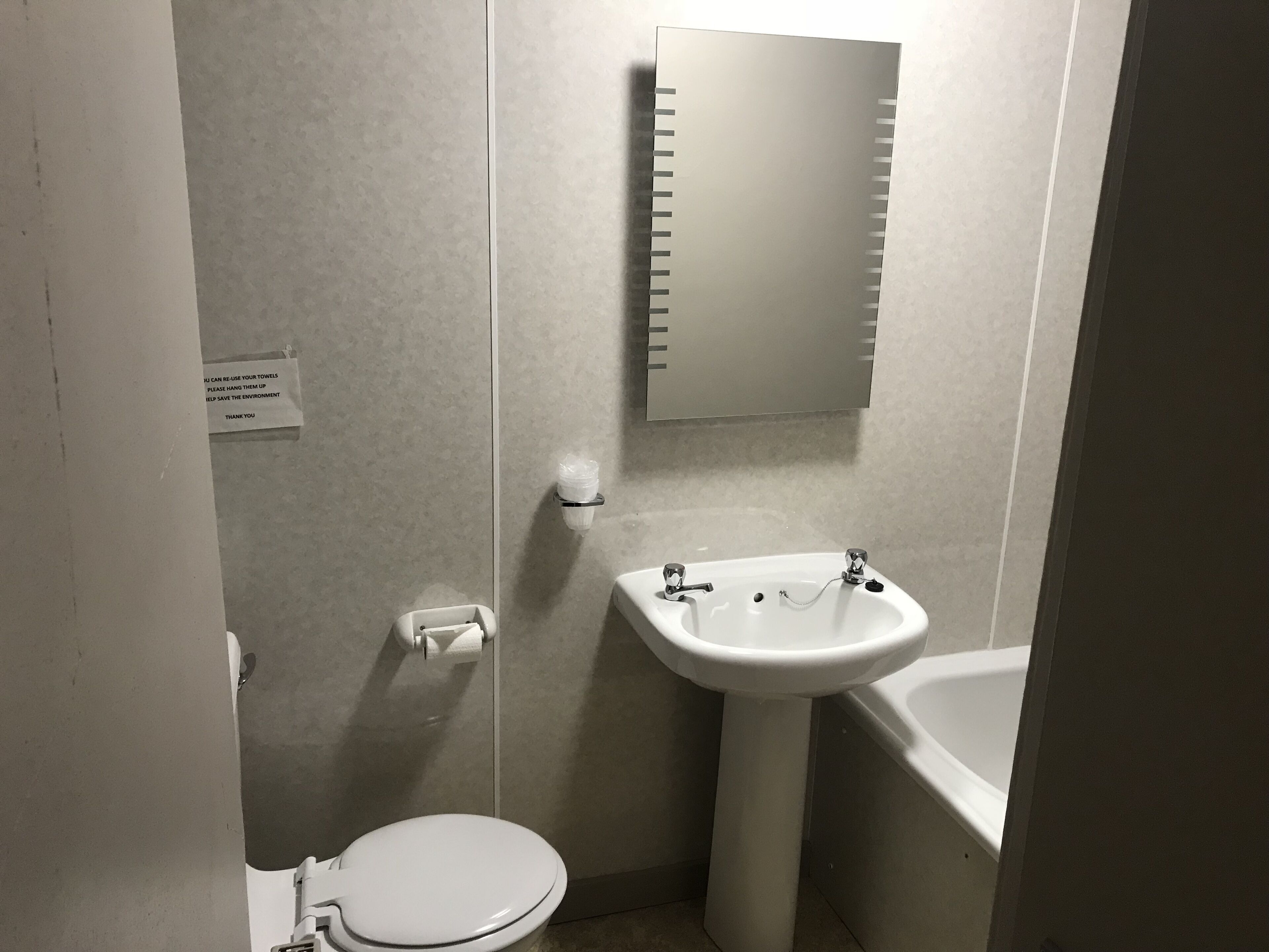 Une petite salle de bain propre, équipée de toilettes, d’un lavabo et d’une baignoire. Un panneau est accroché au mur et un miroir se trouve au-dessus du lavabo.