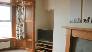 A living room with a fireplace, a television on a wooden stand, and a window with a floral curtain. - House by the Harbour - Nc500 Route (Wick)