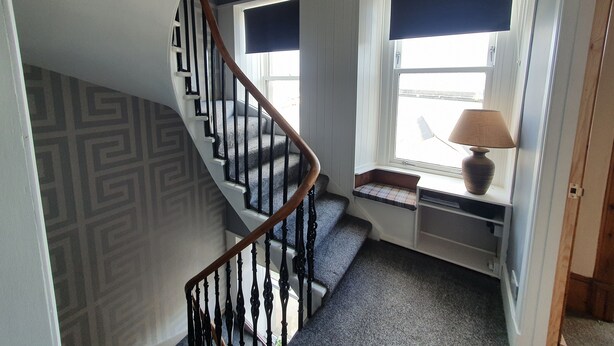 A staircase with a wooden handrail and black balusters, leading to a landing with a window and a lamp. - House by the Harbour - Nc500 Route (Wick)