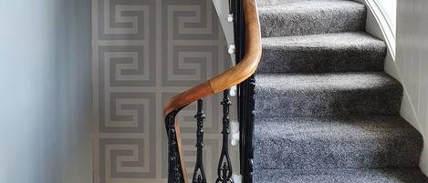 Una escalera de caracol con barandilla de madera y balaustres negros, escalones alfombrados y un papel tapiz decorado.