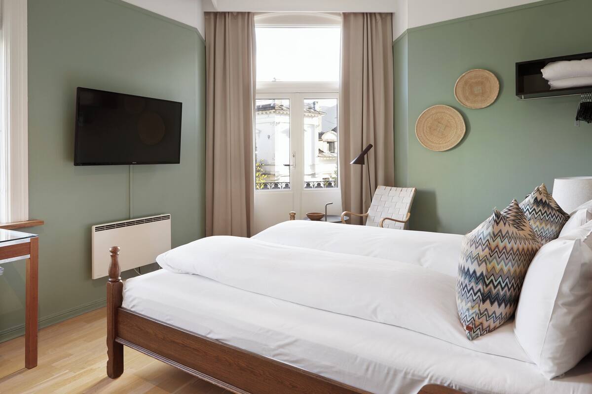 A bedroom with a bed, a TV mounted on the wall, a window with curtains, and a bedside table.