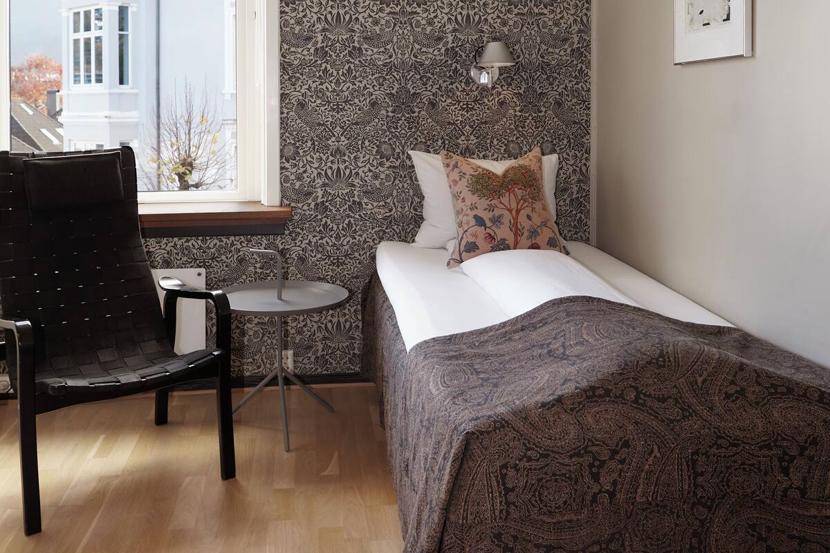 A bedroom with a bed, a chair, a small table, a window, and a patterned wall.