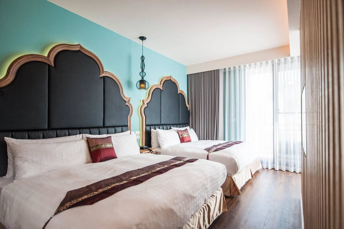 A hotel room with two beds, a large window with curtains, and a modern chandelier.