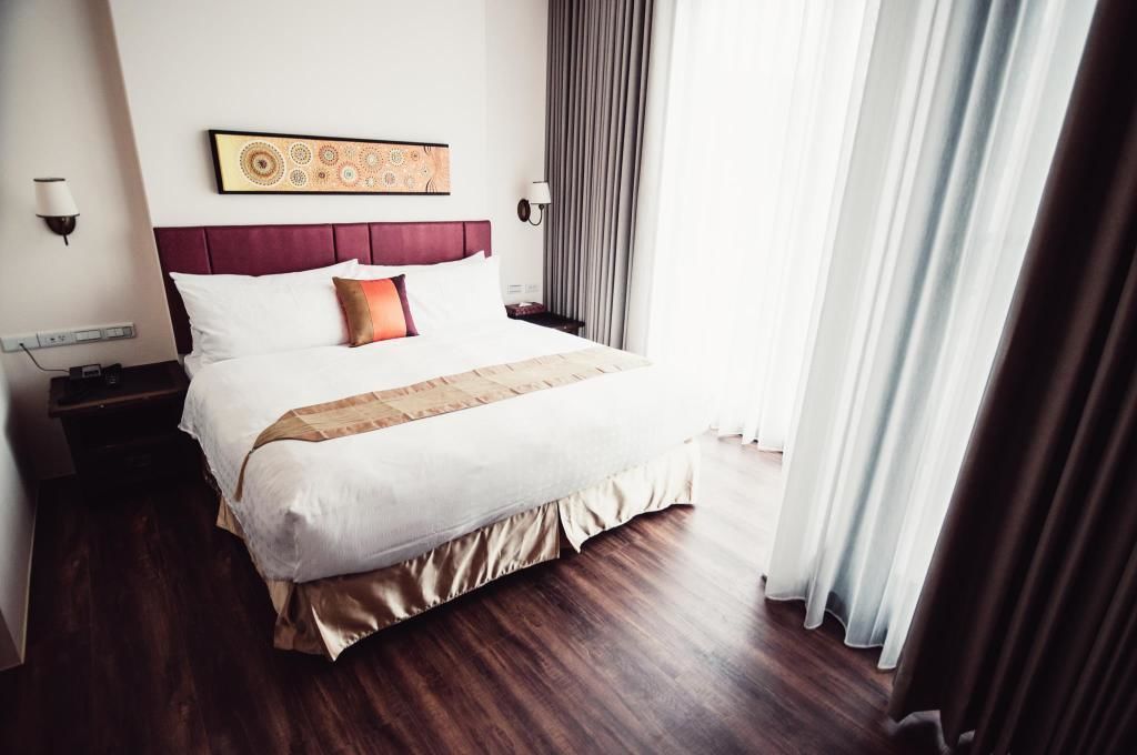 Une chambre d’hôtel avec un grand lit, une tête de lit ornée d’un panneau décoratif, une table de chevet avec une lampe et une fenêtre avec des rideaux.