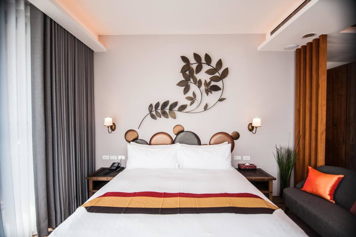 A hotel room with a bed, a headboard with a leaf design, a nightstand, a lamp, and a sofa with an orange pillow.