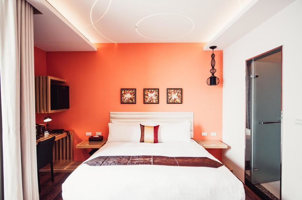A hotel room with an orange wall, a bed with white bedding, a desk with a lamp, and a TV on the wall.