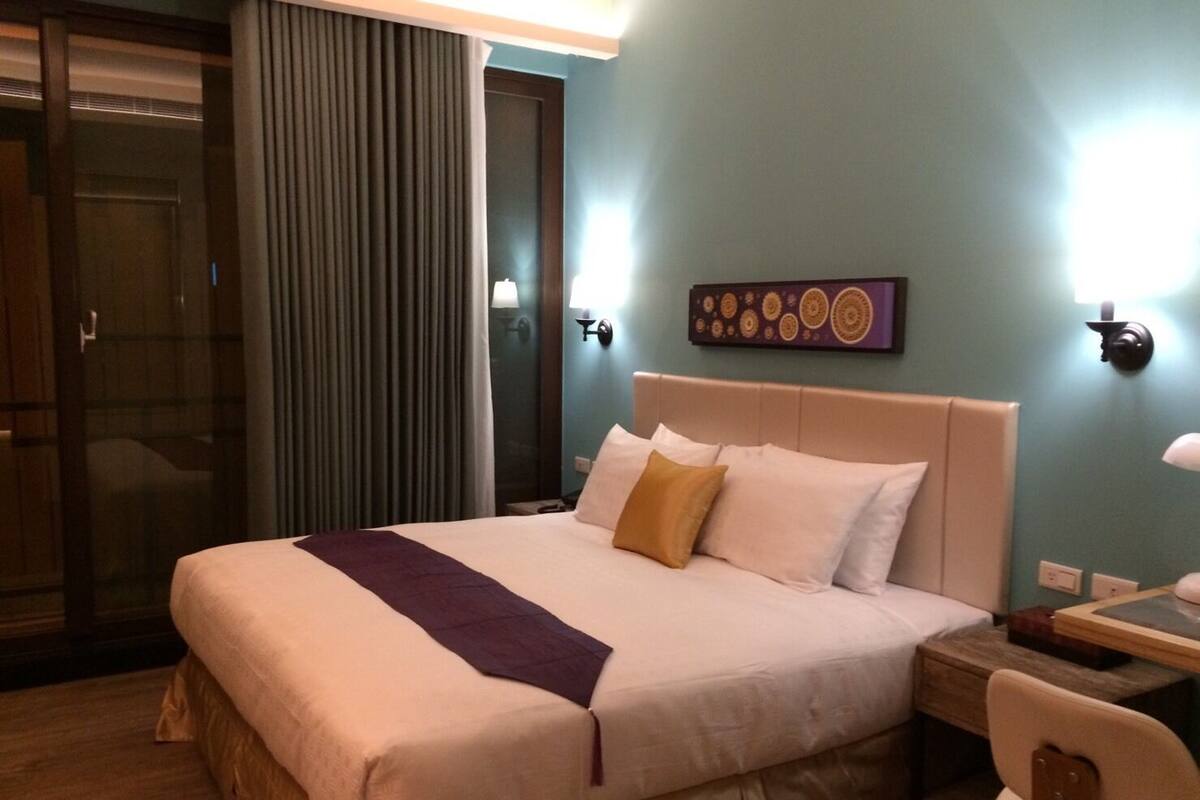 A hotel room with a large bed, a wooden nightstand, a desk with a chair, a sliding glass door, and a wall-mounted artwork.