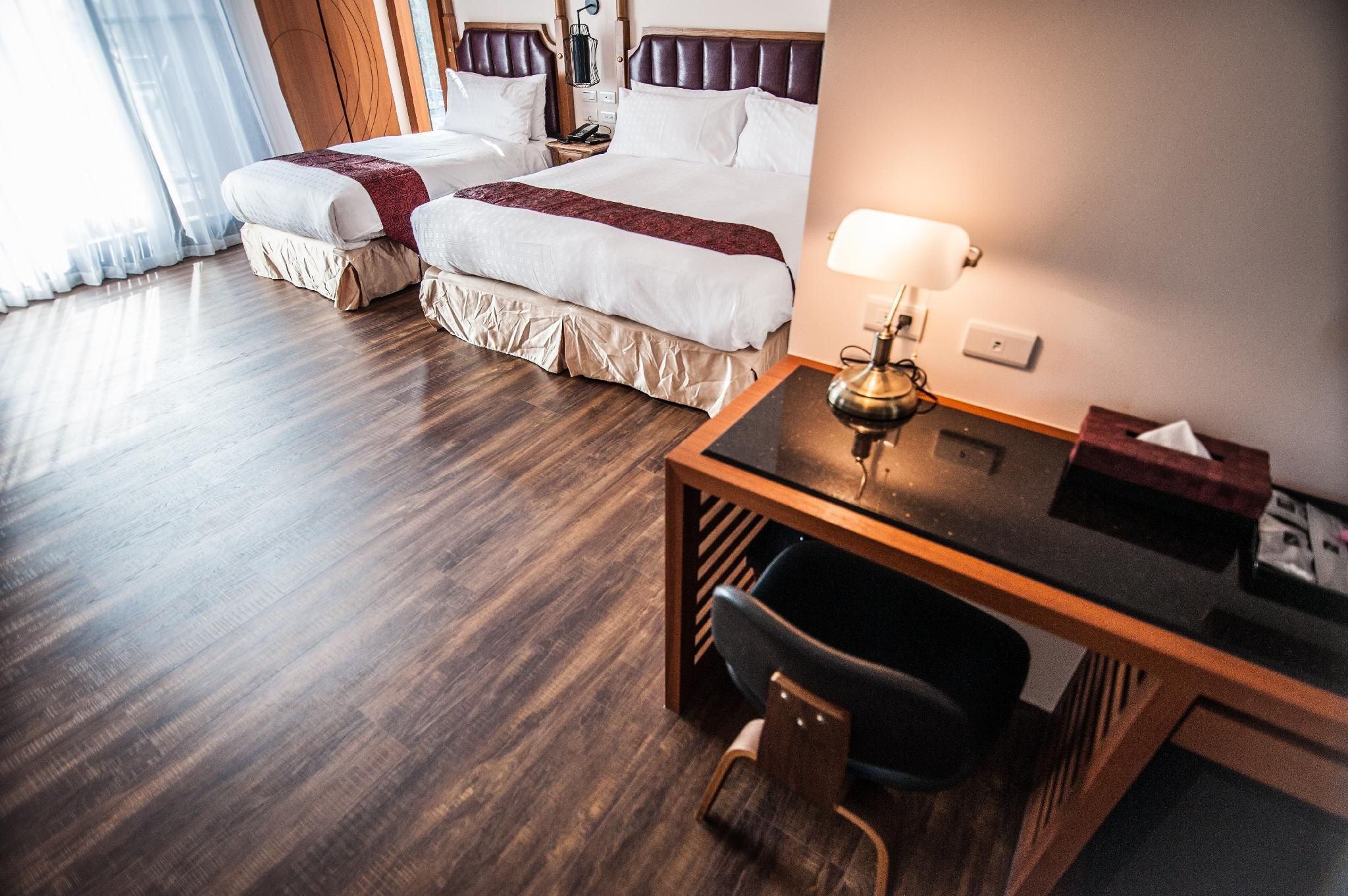 A hotel room with two beds, a desk, a chair, and a lamp.