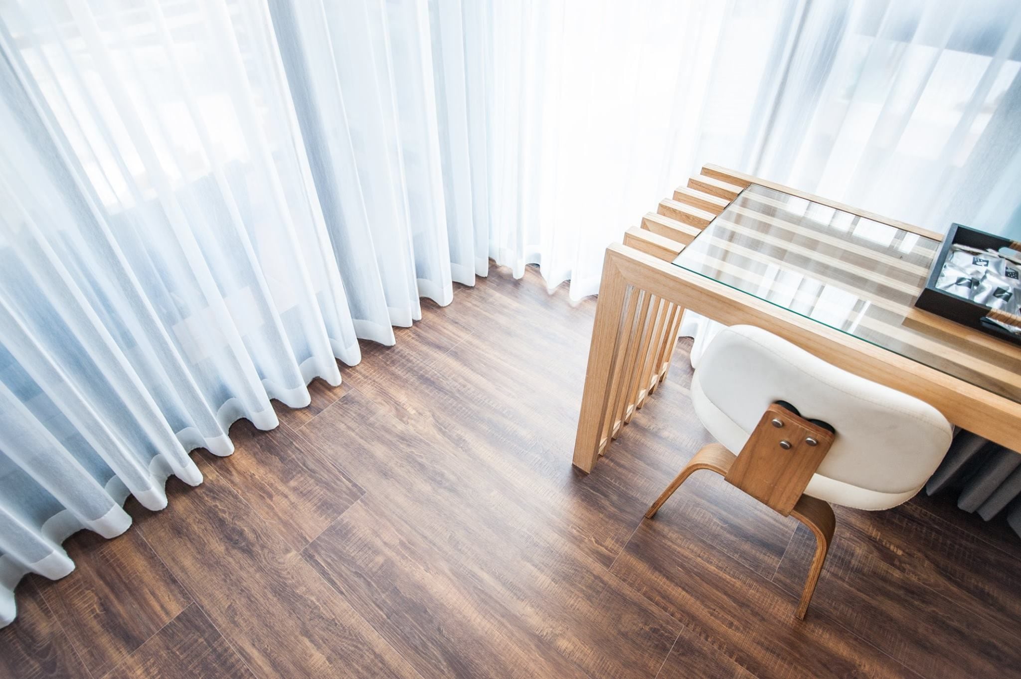 A modern room with a wooden floor, a glass-top table, a white chair, and sheer white curtains.