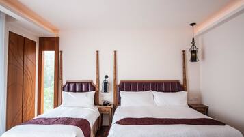 Une chambre double avec deux lits, des têtes de lit en bois, des tables de chevet, une petite table, une lampe et une vue sur la verdure par la fenêtre.