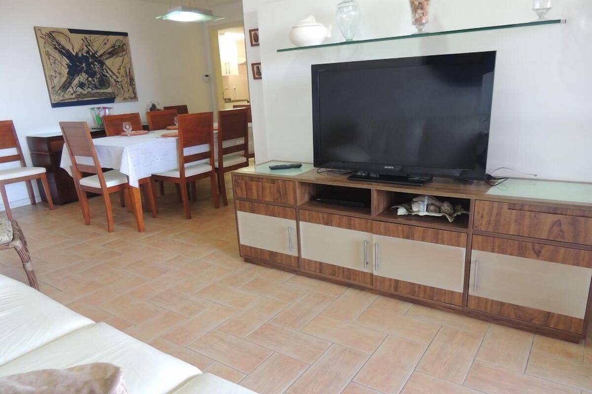 A living room with a TV on a wooden cabinet, a dining area with chairs and a table, and a bed with a blanket.
