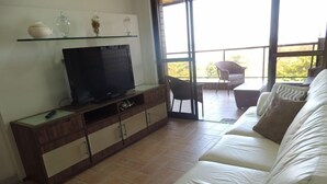 Ein Wohnzimmer mit Fernseher auf einer Holzeinrichtung, einem weißen Sofa und einem Balkon mit Stühlen.
