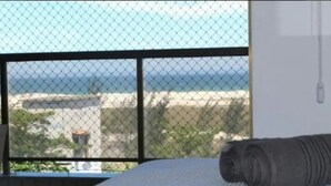 Ein Schlafzimmer mit einem Bett, einem Fenster mit Blick auf Gebäude und einen Zaun, sowie einer zusammengerollten Decke auf dem Bett.