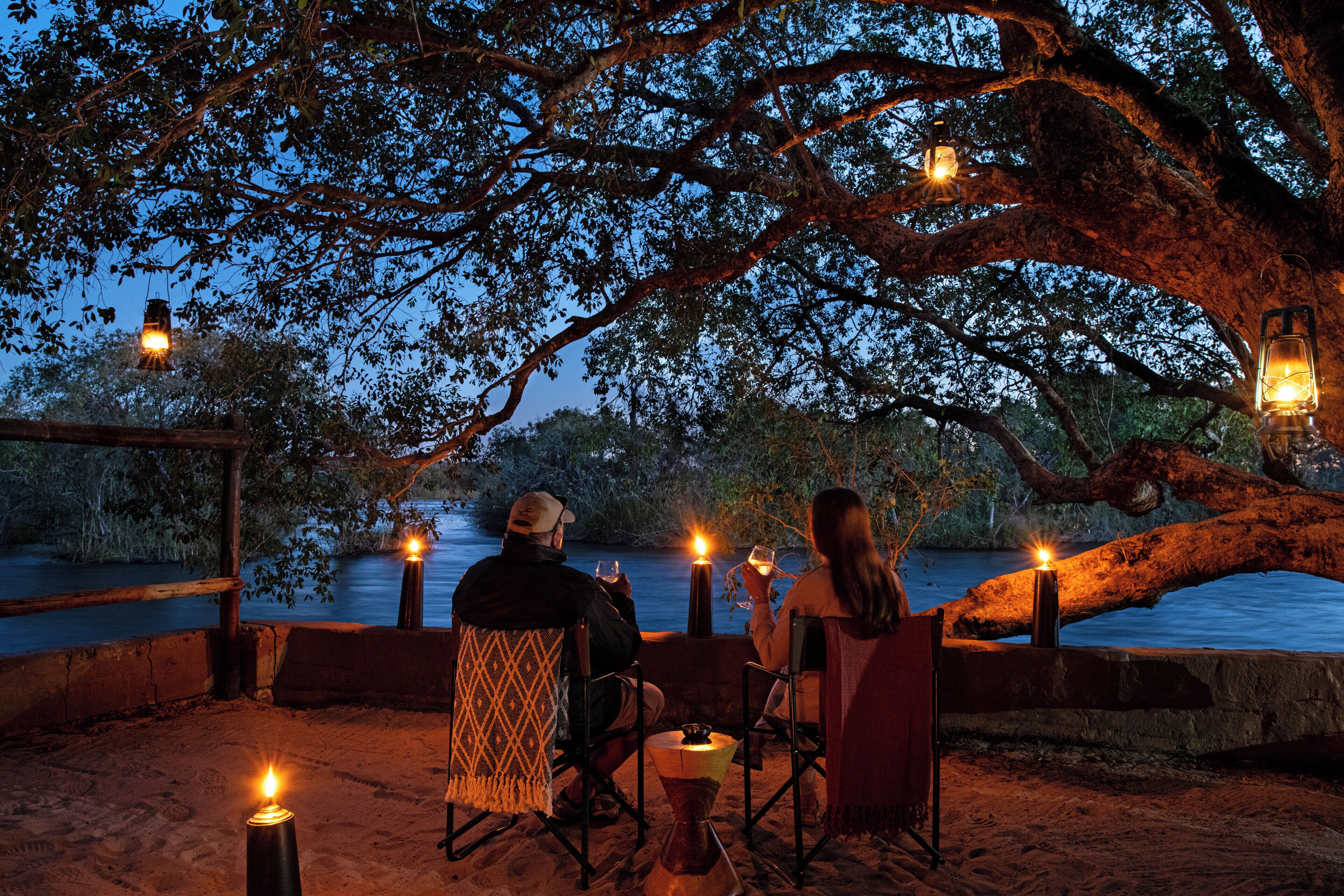 A couple sitting on chairs by a lakeside at dusk, with candles and lanterns providing light.