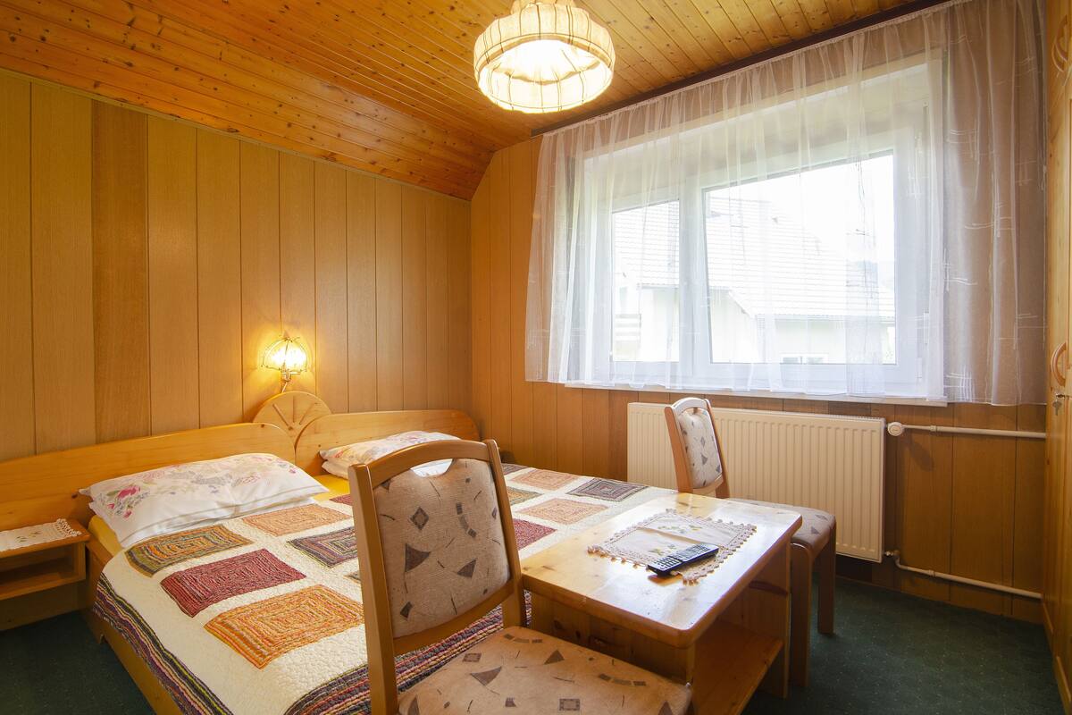 A cozy room with a wooden ceiling, a bed with a quilt, a table with a chair, a window with sheer curtains, and a wall-mounted lamp.