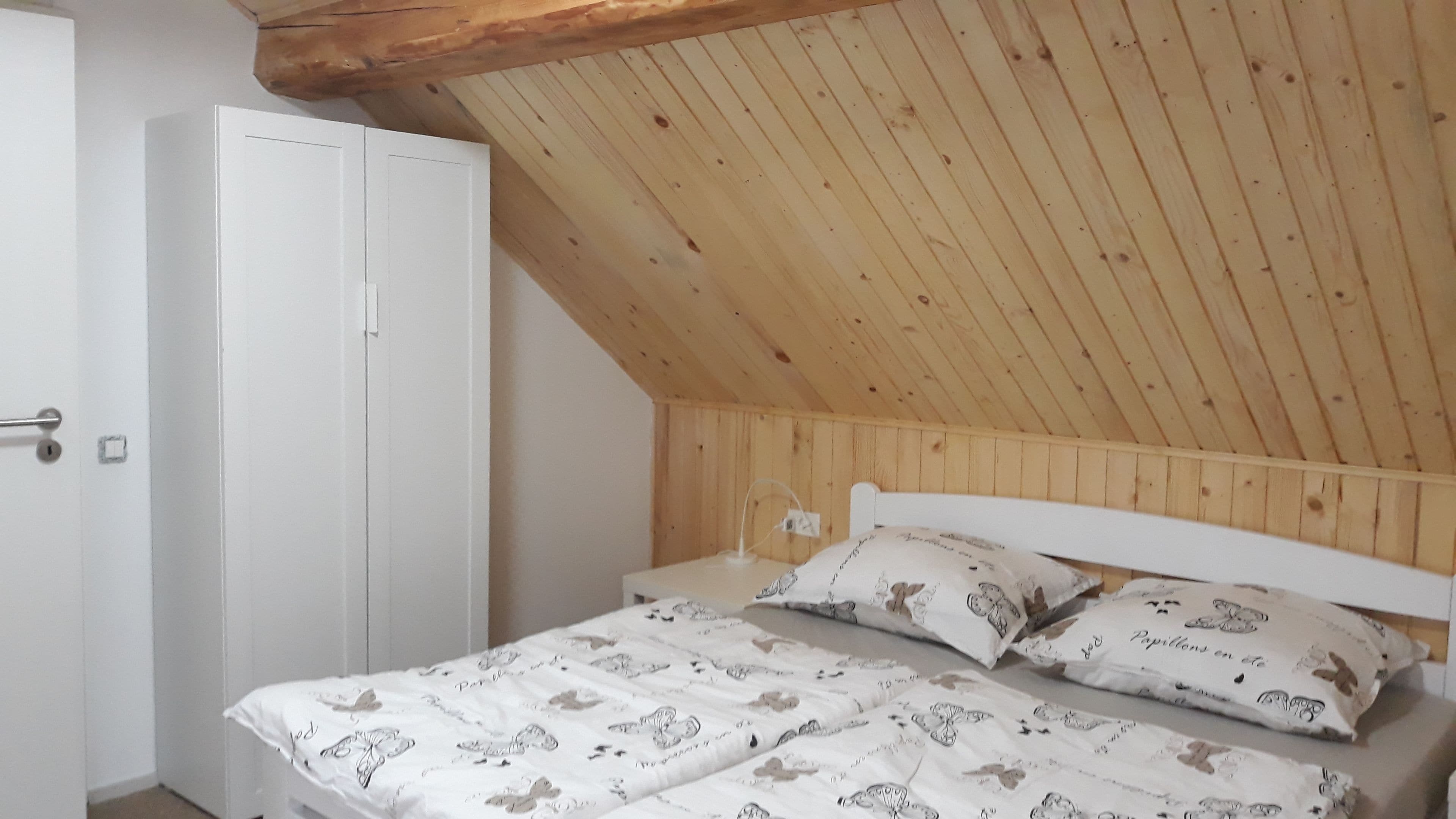 Una camera da letto con soffitto in legno inclinato, un letto bianco con cuscini decorati e un armadio bianco.