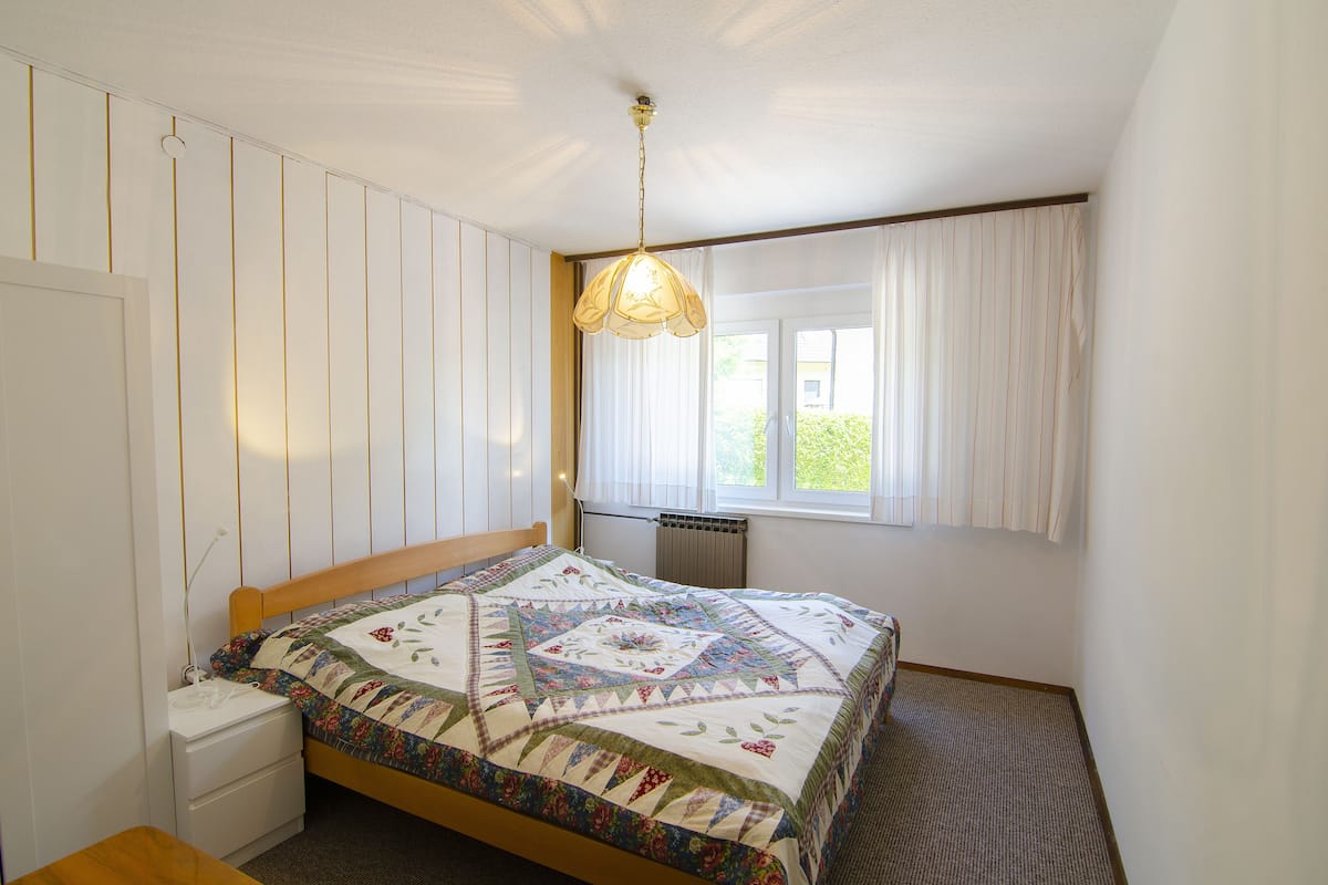 A bedroom with a wooden bed, a nightstand, a window with curtains, and a hanging light fixture.