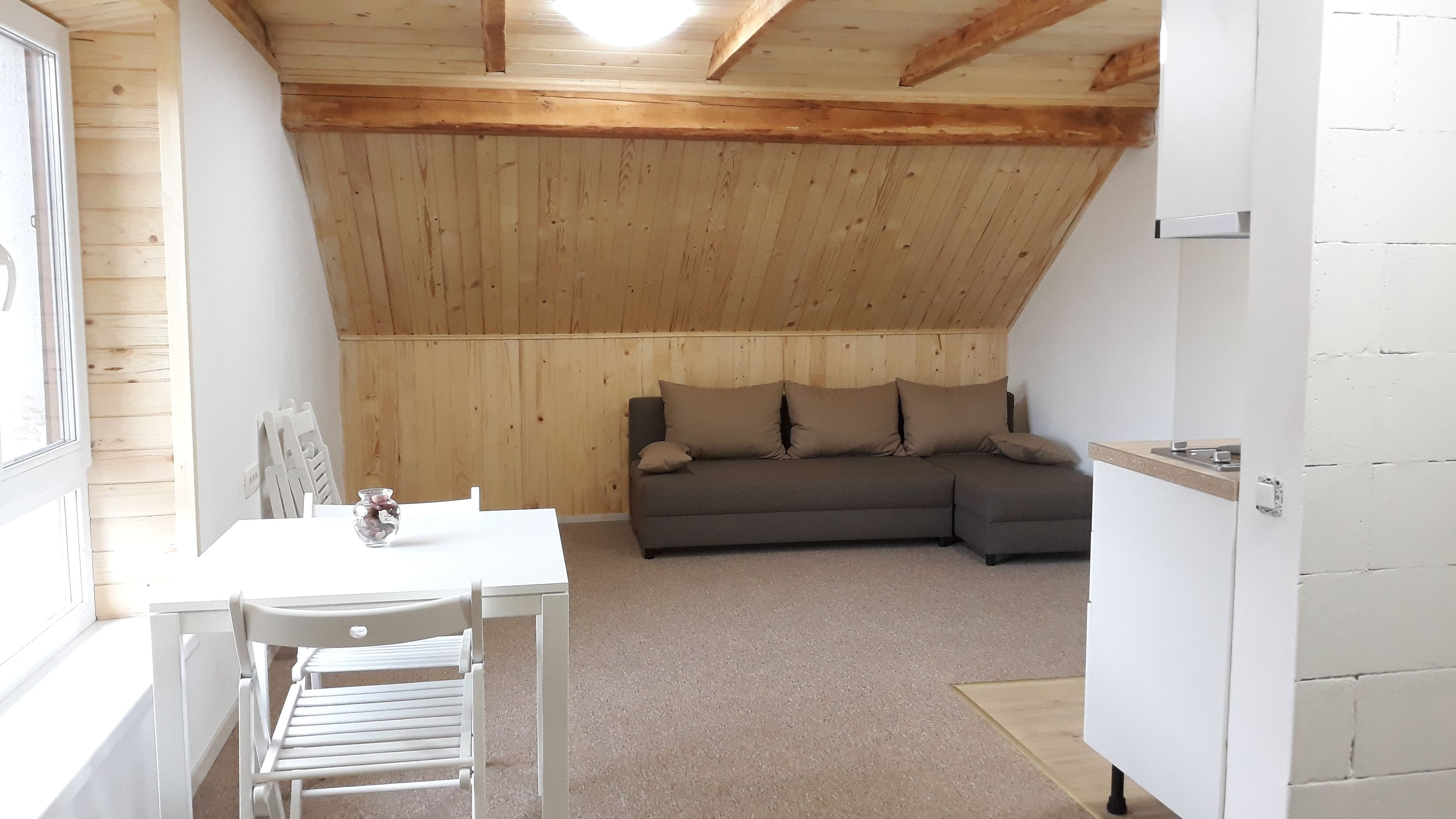 Ein gemütliches Schlafzimmer unter dem Dach mit Holzbalkendecke, einem grauen Sofa mit Kissen, einem kleinen Tisch mit einer Vase und einem weißen Sessel.