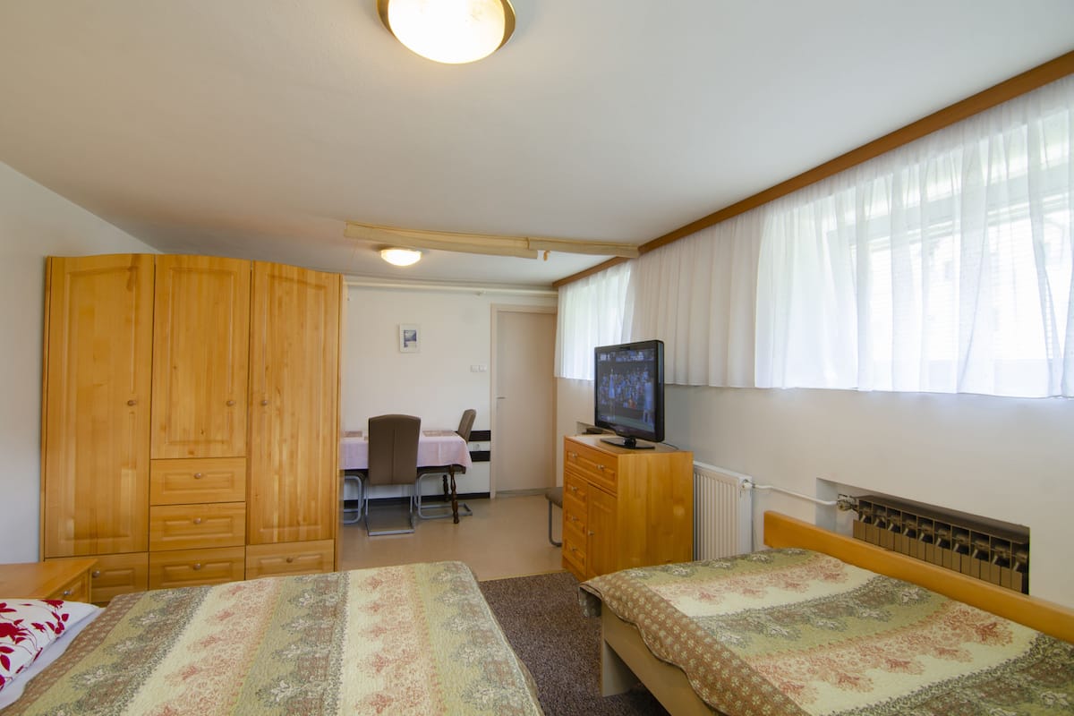 A room with two beds, a wooden wardrobe, a television on a wooden cabinet, a desk with a chair, and a window with curtains.