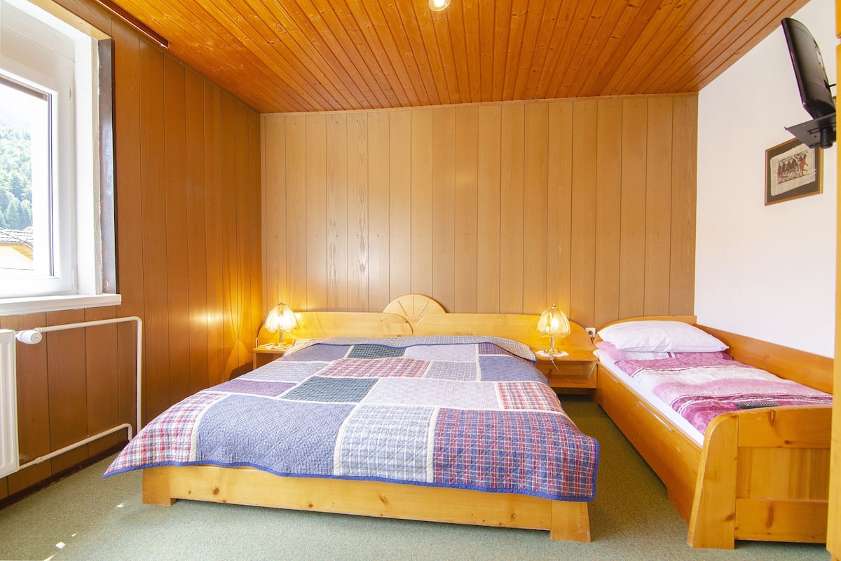 A room with a wooden ceiling, a large bed with a quilt, two bedside tables with lamps, and a window with a view of trees.