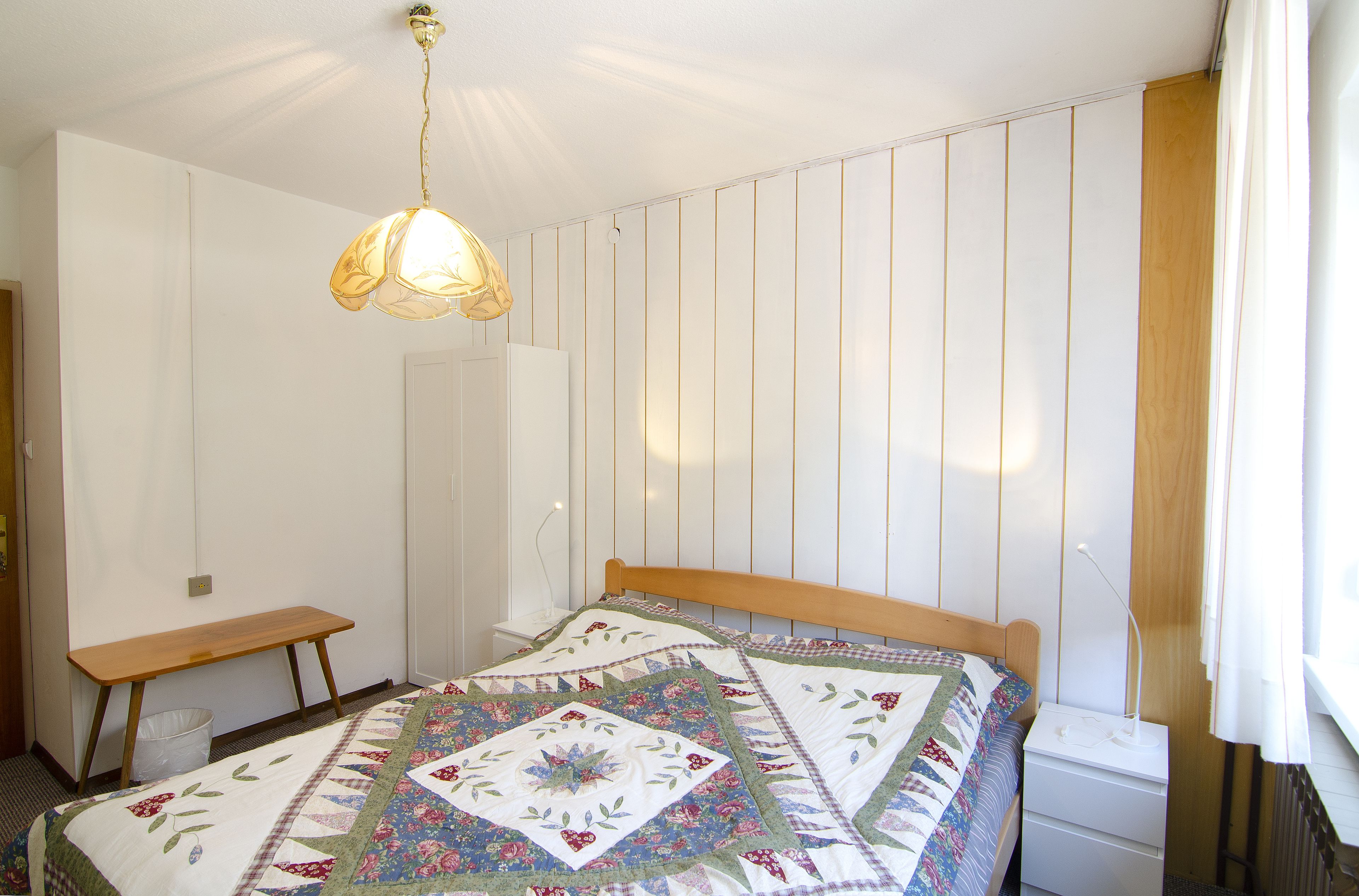 A bedroom with a wooden bed, a bedside table, a nightstand, a hanging light fixture, and a window with curtains.
