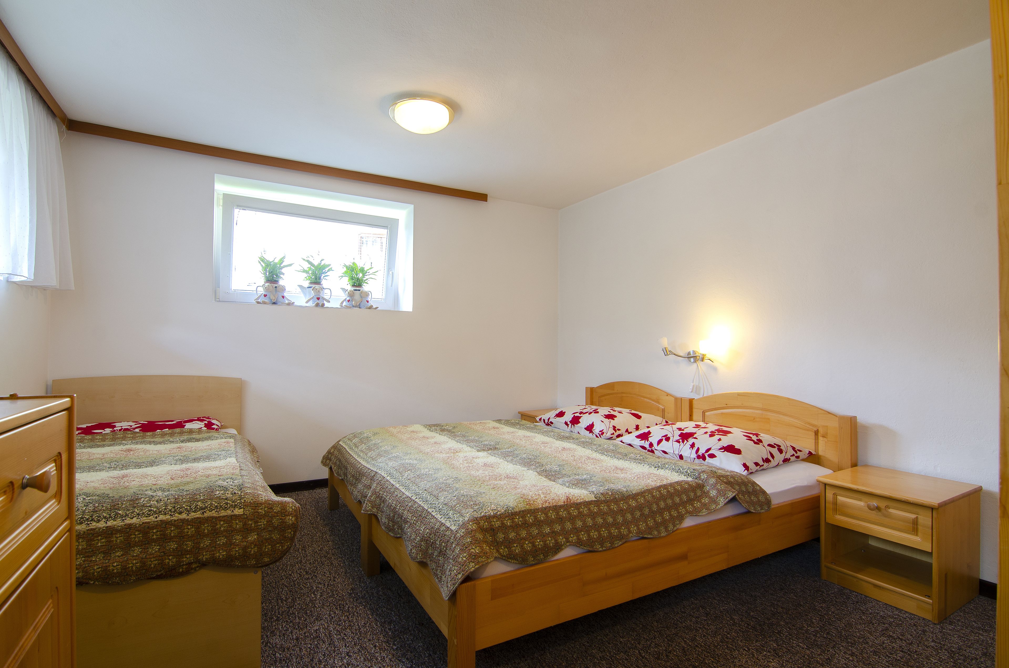 A bedroom with two single beds, a window with potted plants, a bedside table with a lamp, and a wooden dresser.