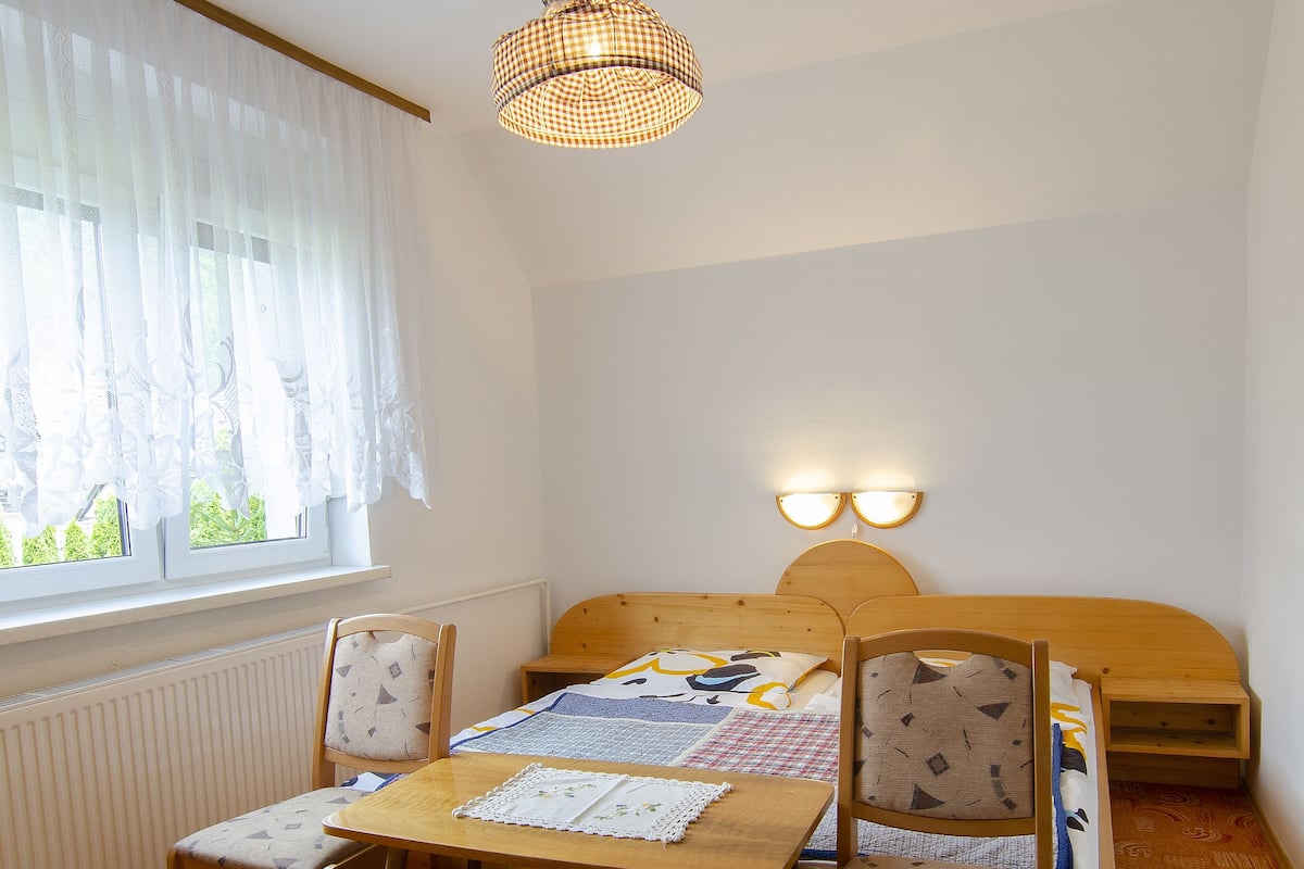 A room with a wooden table and chairs, a bed with a headboard, a window with sheer curtains, and a hanging light fixture.