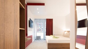 Habitación de hotel con cama, televisor montado en la pared, armario de madera y ventana con cortinas.