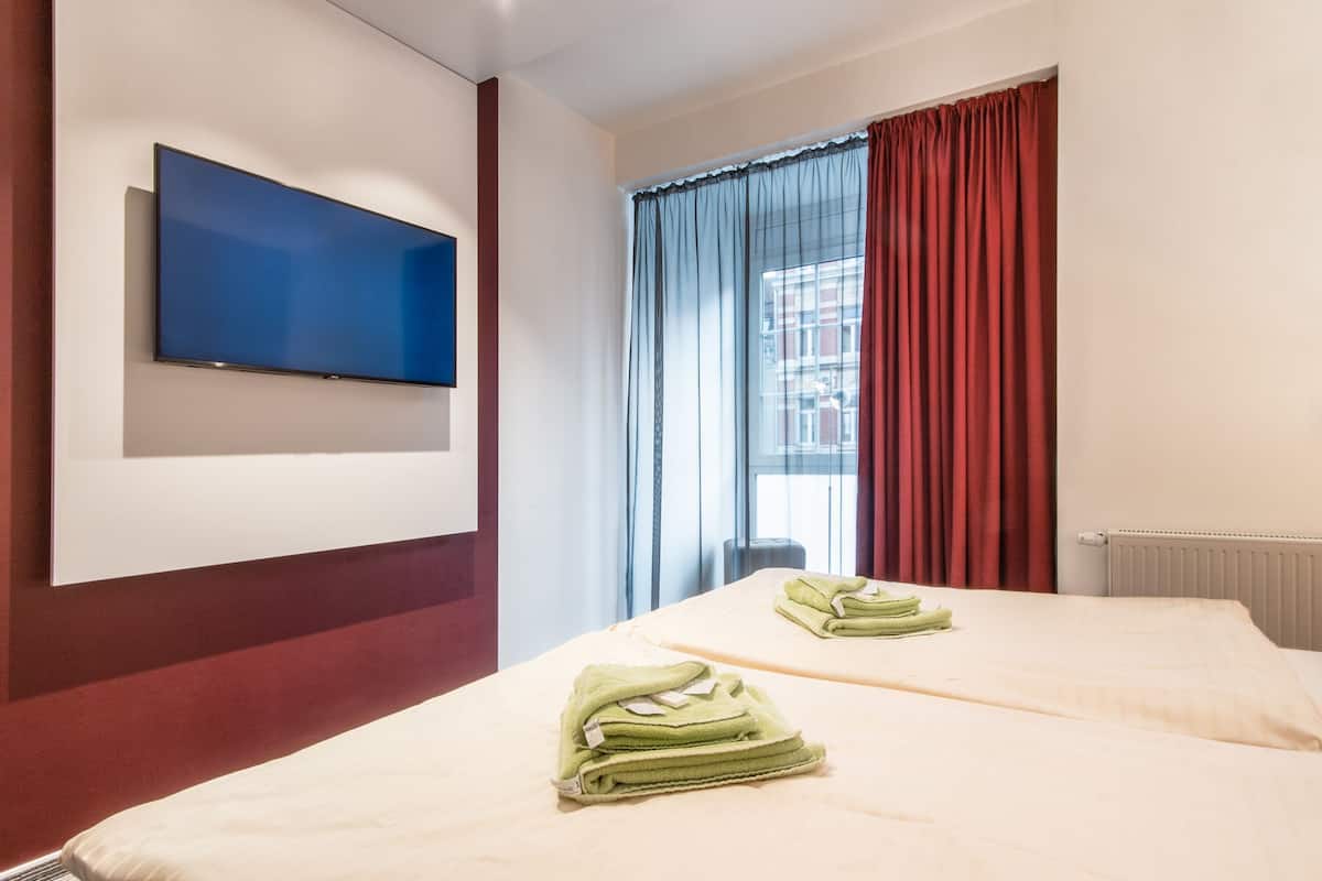 A hotel room with a flat-screen TV mounted on the wall, two beds with neatly folded towels, a window with red curtains, and a radiator.