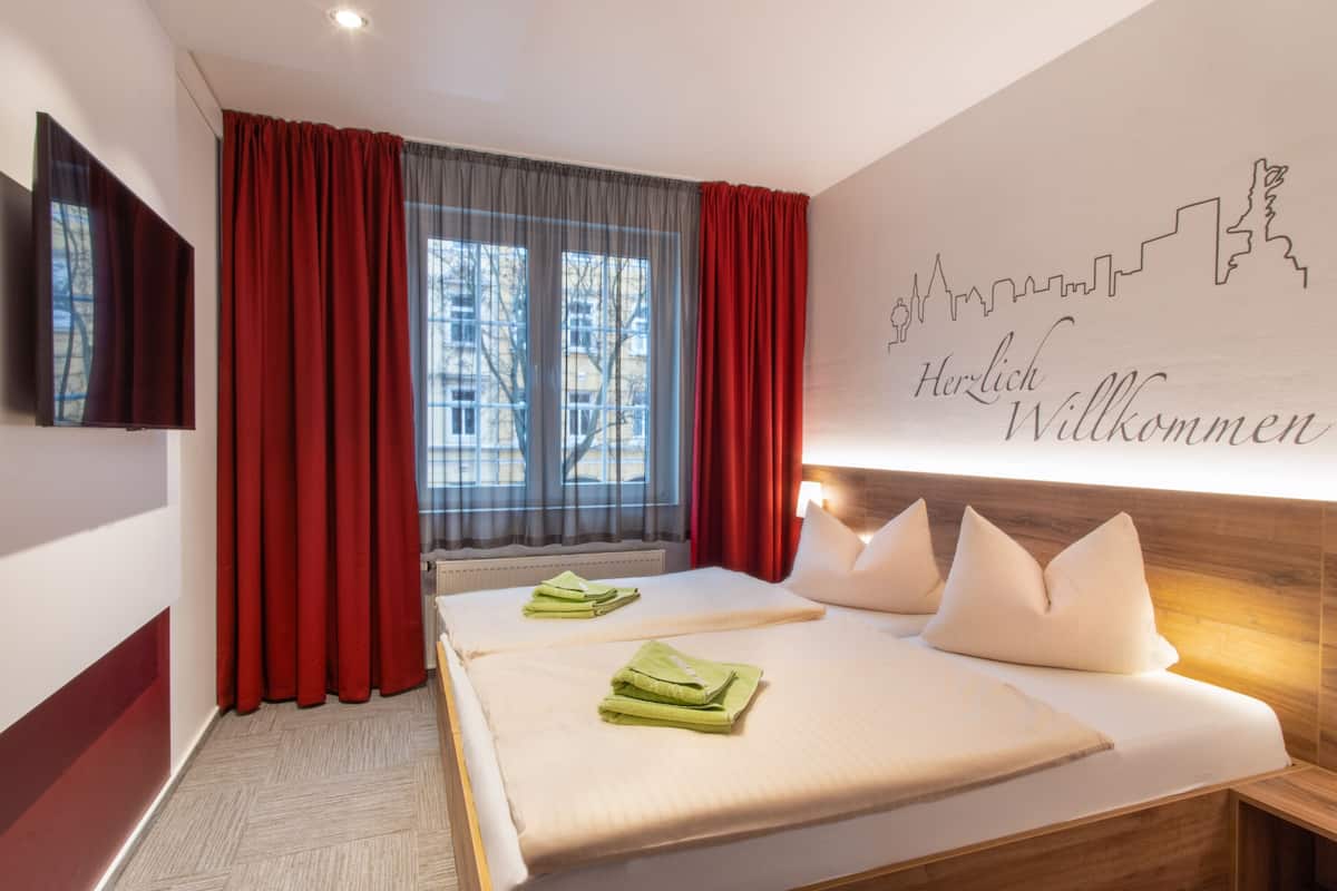 A hotel room with a large bed, red curtains, a city skyline wall art, and a flat-screen TV on the wall.