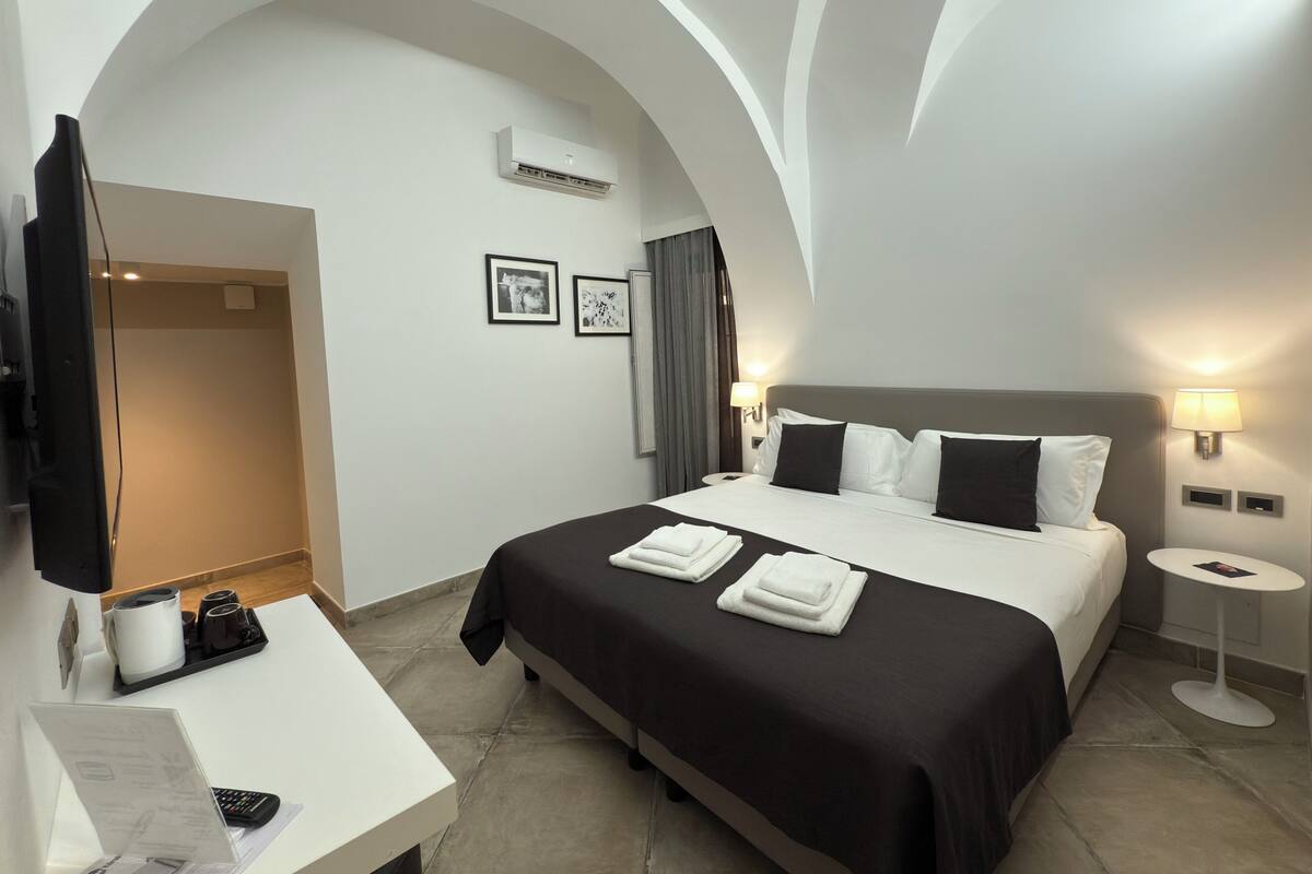 A hotel room with a bed, a desk, a flat-screen TV, a coffee machine, and a wall-mounted air conditioner.