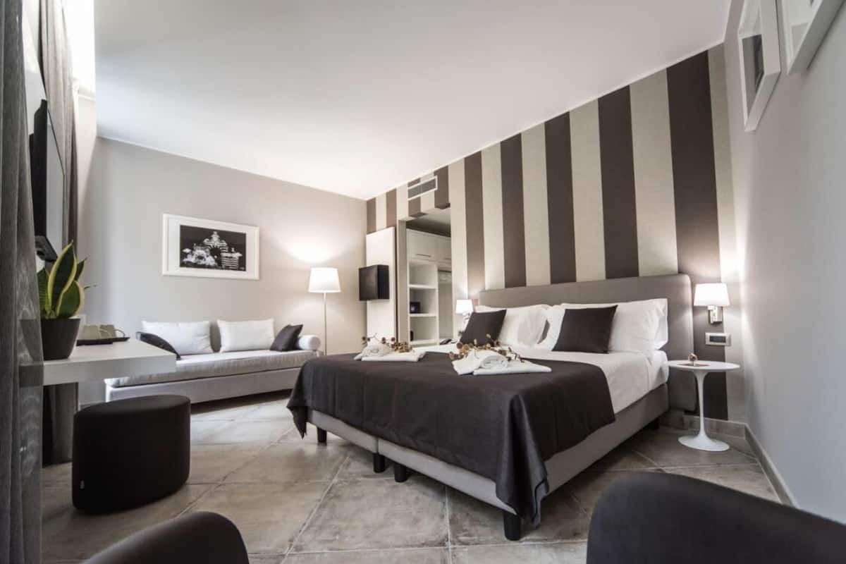 A modern hotel room with a large bed, a desk, and a chair. There is a mirror on the wall and a plant on the desk.