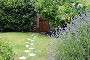 Un jardin avec un chemin de pierres menant à une porte en bois, entouré d’une végétation luxuriante et de plantes de lavande.