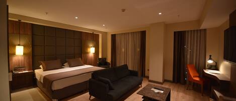 A hotel room with a large bed, a sofa, a desk, and a chair. There are two lamps on the left side of the bed and one lamp on the desk.