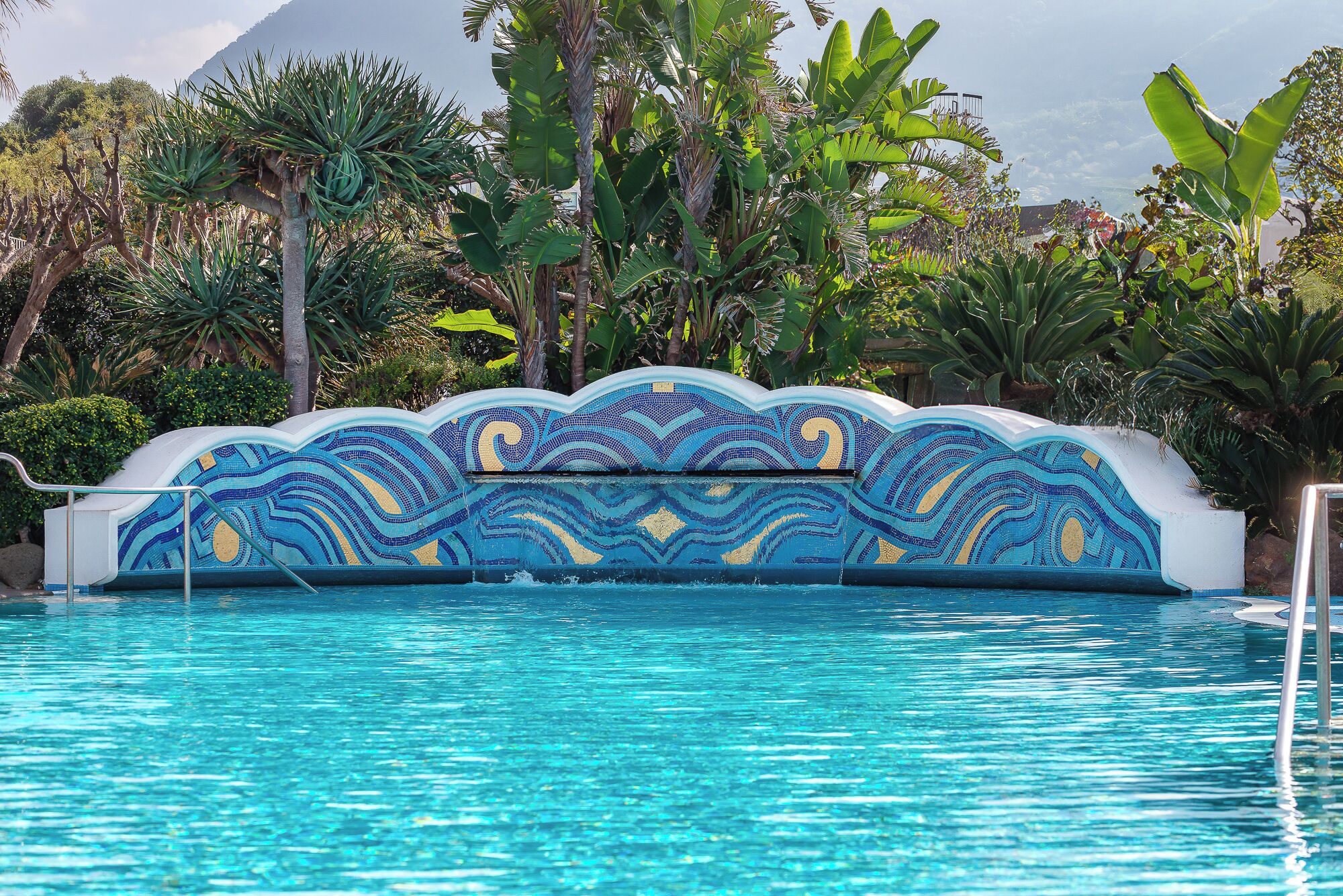 A swimming pool with a mosaic tile wall and tropical plants around it.
