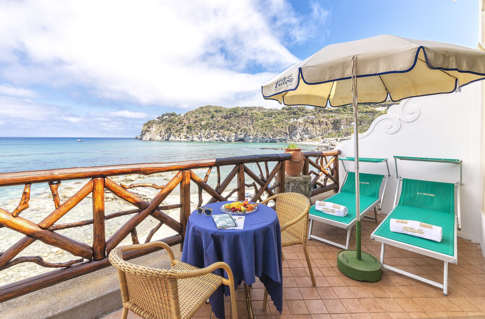 A terrace with a table, chairs, and a lounge chair, overlooking a beach and the sea.