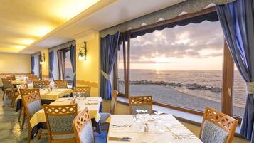 A dining area with tables set for four, overlooking a beach with a sunset view.